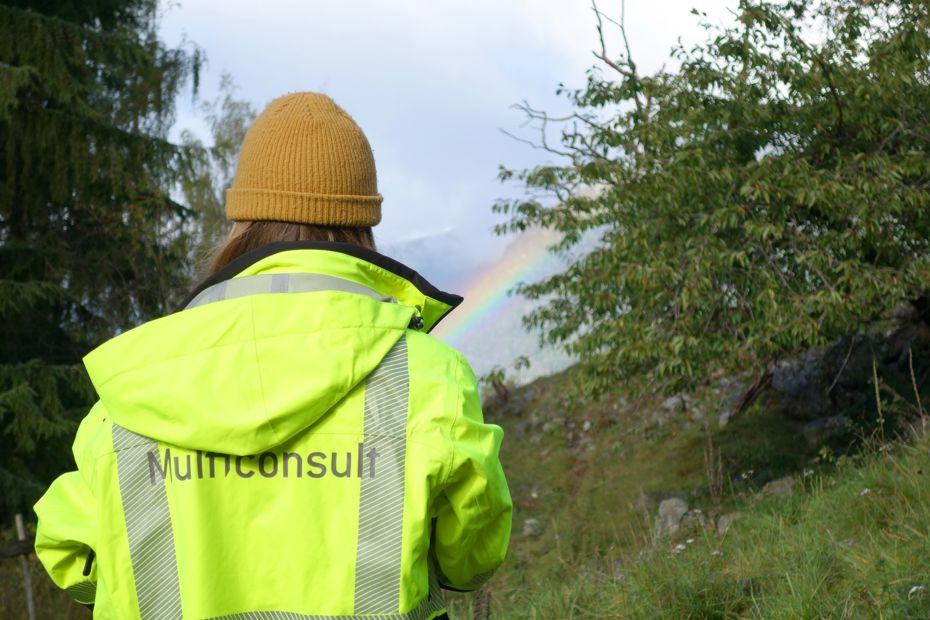 Kvinne sett bakfra, ikledd vernetøy med Multiconsultlogo på rygg, ute i naturen.